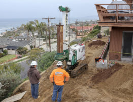 Two men in hardhats observe a small access drill on a hill overlooking houses and shoreline in La Jolla, CA.