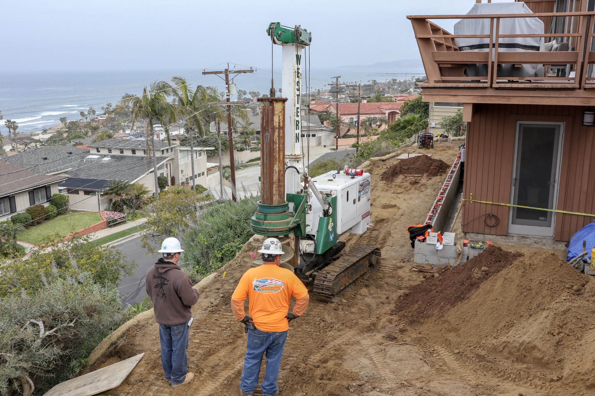 Two men in hardhats observe a small access drill on a hill overlooking houses and shoreline in La Jolla, CA.