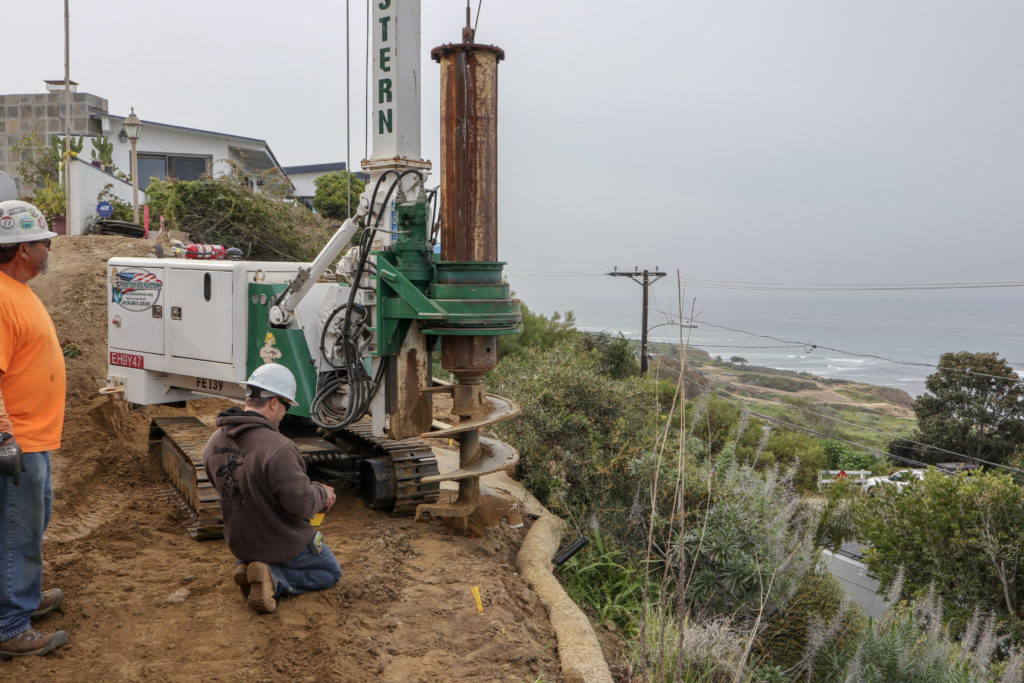 Two construction workers use a limited-access drill rig to dig on the edge of a hill in this residential project in La Jolla.
