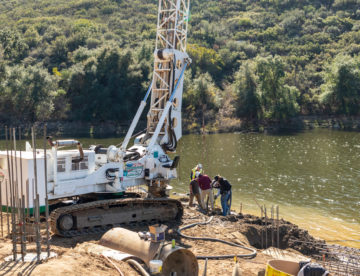 A drill rig overlooks a lake, ready to create caissons for a lakeside swimming pool as men work on a nearby caisson.
