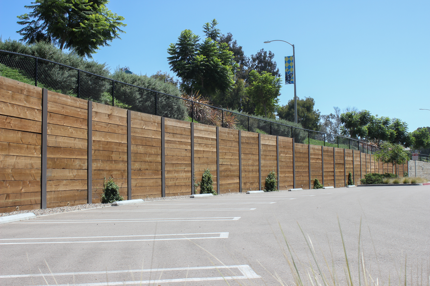 A custom wooden soldier pile shoring in a parking lot acts as a rustic retaining wall with bushes and trees peeking above.